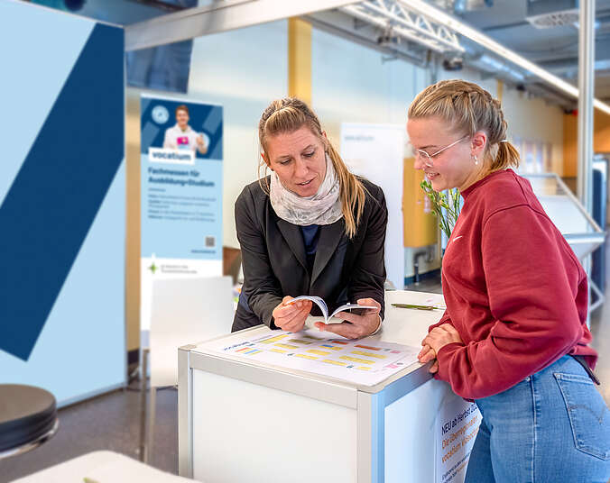 Eine junge Frau und eine Frau mittleren Alters unterhalten sich an einem Messestand und schauen zusammen in ein Smartphone.