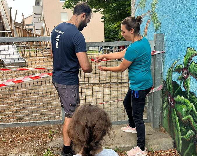Wieder geöffnet: Mann und Frau zerschneiden Band an Spielplatztüre, Kind in Rückansicht wartet