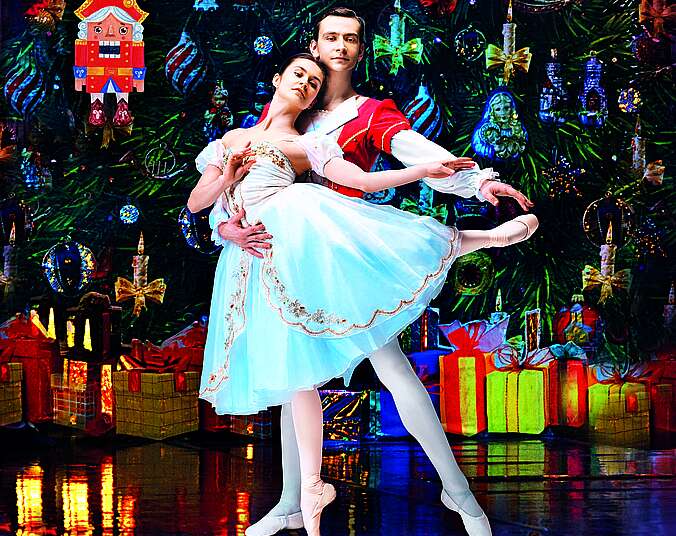 Eine junge Ballettt-Tänzerin in zartem hellblauem Kleid und ein junger Ballett-Tänzer in roter Uniformjacke stehe vor einem riesengroßen, geschmückten Weihnachtsbaum. Links von ihnen hängt ein großer Nussknacker im Baum.