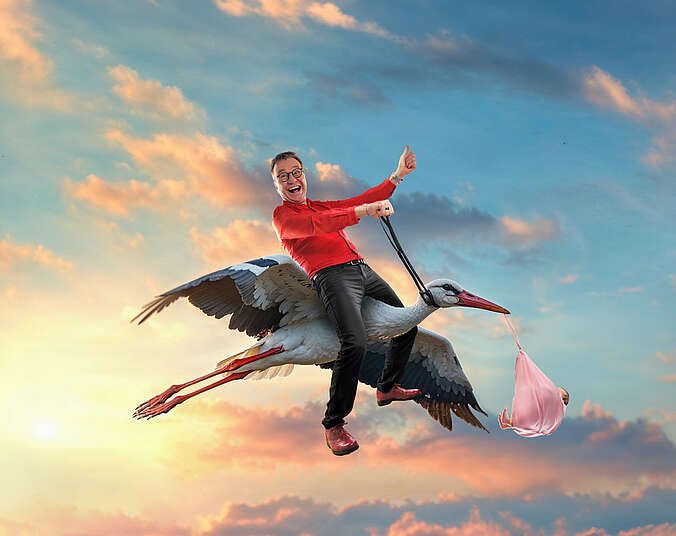 Ein Mann um die 50 reitet auf einem fliegenden Storch durch die Lüfte. Er trägt ein rotes Hemd und dunkle Jeans. Der Storch trägt ein rosa Windelpaket mit einem Baby im Schnabel.tuch