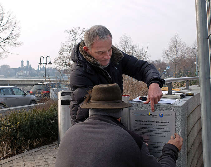 Stefan Fehringer (links) und Reinhard Polensky (rechts) vom städtischen Amt für Vermessung und Liegenschaften haben an der Mauer zwischen Partnerschaftsgarten und dem Eingang zum Parkhaus am See den Kontrollpunkt angeschraubt.