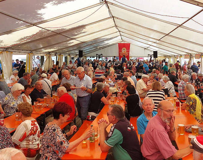 Menschen sitzen auf Bierbänken an Biertischen in einem Zelt