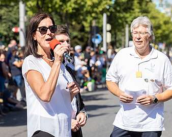Festzug: Frau und Mann moderieren auf Straße, Frau spricht in Mikrofon