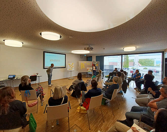 Mehrere Personen sitzen auf Stühlen, Moderator hält Vortrag.