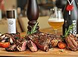 Aufgeschnittenes Rindfleisch, innen rosa mit Kräutern und Tomate auf einem Holzbrett. Im Hintergrund ein Glas Bier.