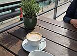 Eine Tasse Kaffee und ein Dessert stehen auf einem Tisch, im Hintergrund ist der Hafen von Friederichshafen mit dem Moleturm zu sehen.